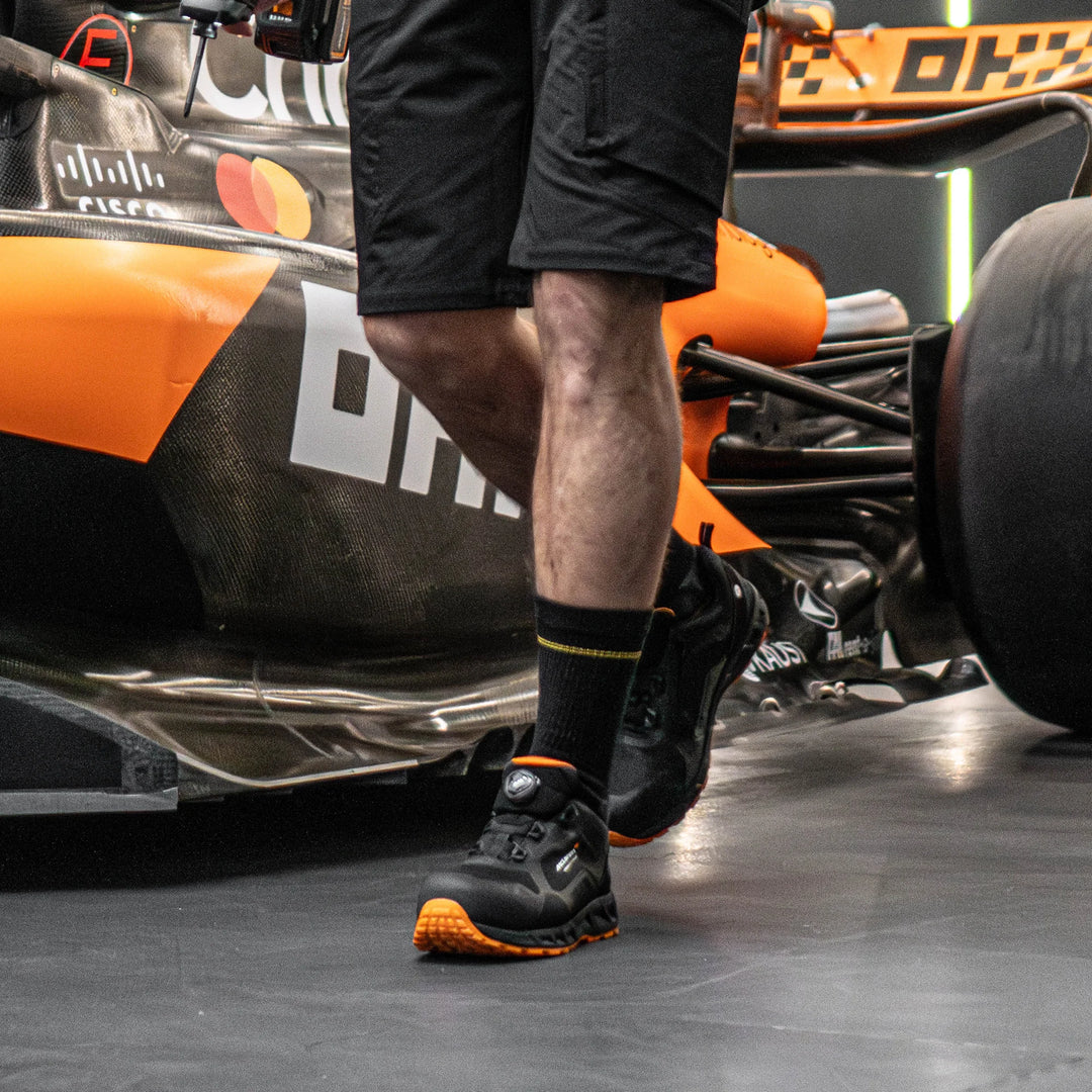 Person wearing black shoes with orange accents standing next to a Formula 1 car.