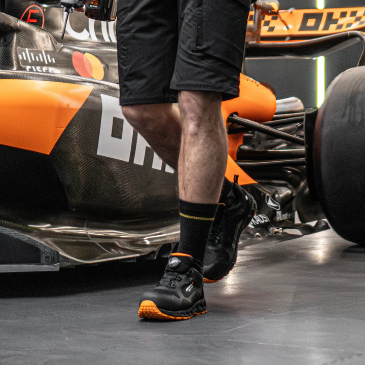 Person wearing black shoes with orange accents standing next to a Formula 1 car.